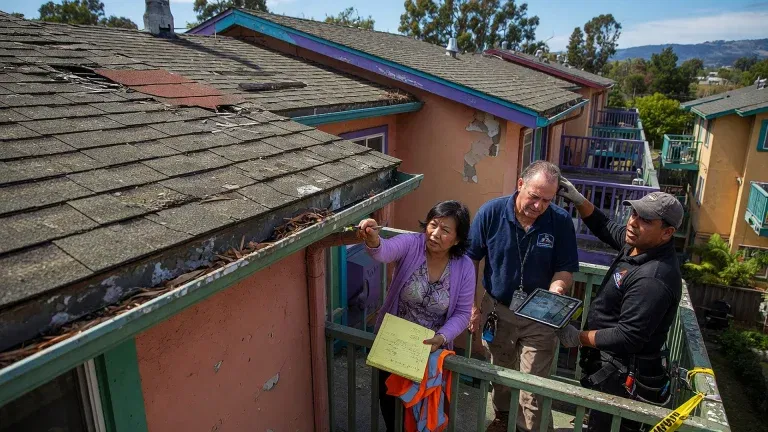 Three people inspect the exterior of an old apartment building with damaged roofs and peeling paint. As they discuss needed repairs, East Bay HOAs consider future expenses and reserve fund management to address maintenance issues.