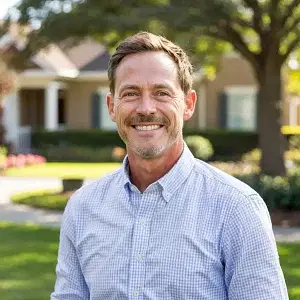 A smiling man with short brown hair and a beard is standing outdoors. He wears a light checkered shirt. The background shows a suburban neighborhood with a house, trees, and greenery under a clear sky.