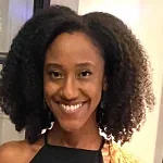 A woman with natural, curly hair smiles at the camera. She is wearing a black halter top and is standing indoors in front of a window and white wall.