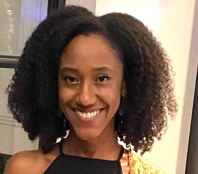 A woman with natural, curly hair smiles at the camera. She is wearing a black halter top and is standing indoors in front of a window and white wall.