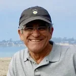 A smiling older man wearing glasses, a black cap, and a gray polo shirt stands outdoors with a blurred background of water and distant land, reflecting the approachable spirit of an experienced Oakland HOA Management team.