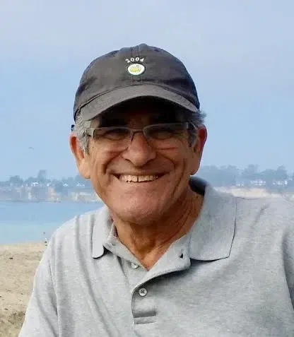 A smiling older man wearing glasses, a black cap, and a gray polo shirt stands outdoors with a blurred background of water and distant land, reflecting the approachable spirit of an experienced Oakland HOA Management team.