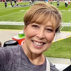 A woman with short, light brown hair and a gray shirt smiles for a selfie at a football stadium, with the field, sideline, and other people visible in the background—capturing the fun spirit of an Oakland HOA management team outing.