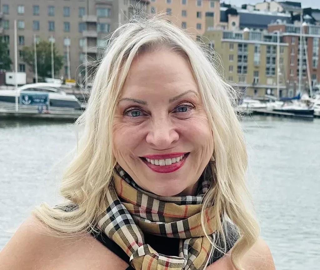 A smiling woman with long blonde hair stands in front of a waterfront with boats and modern apartment buildingsâperhaps the ideal spot for an Oakland HOA management team to oversee. She is wearing a beige, plaid scarf and a black top.