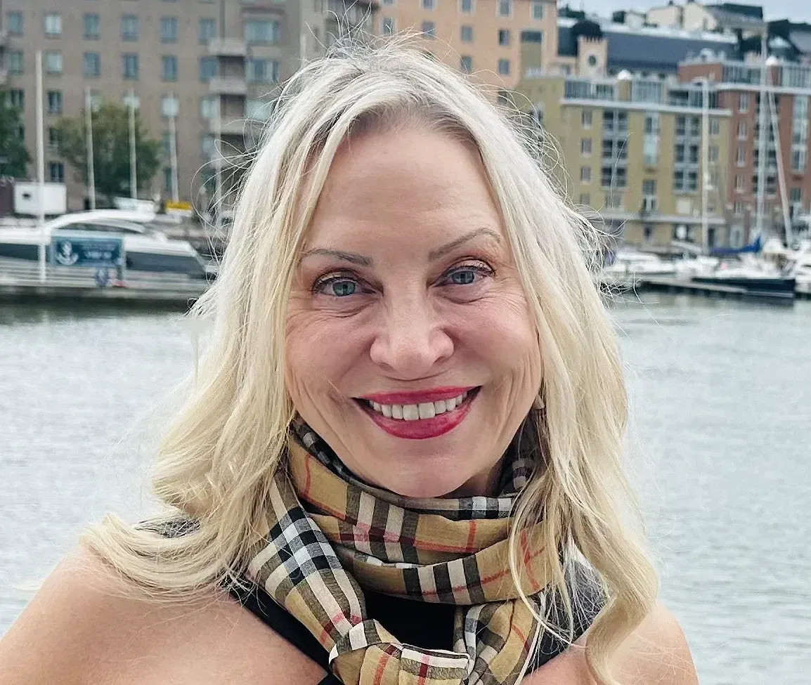 A smiling woman with long blonde hair stands in front of a waterfront with boats and modern apartment buildings—perhaps the ideal spot for an Oakland HOA management team to oversee. She is wearing a beige, plaid scarf and a black top.