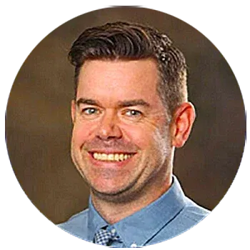 A man with short dark hair smiles at the camera, wearing a blue collared shirt and a patterned tie, against a neutral brown background.