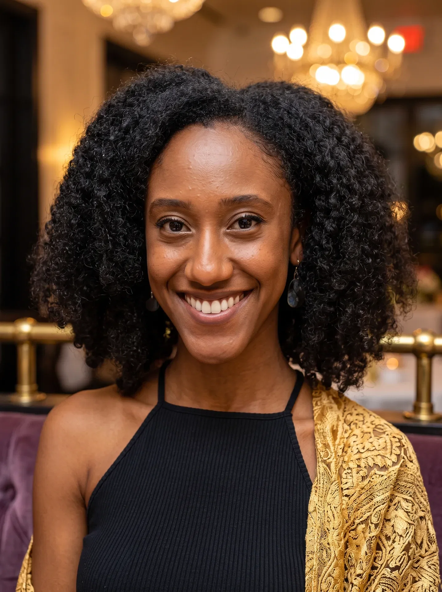 A woman with curly hair and a bright smile wears a black top and gold-patterned shawl, sitting indoors in the San Francisco Bay Area, with chandeliers glowing aboveâperfect for business association management services events.