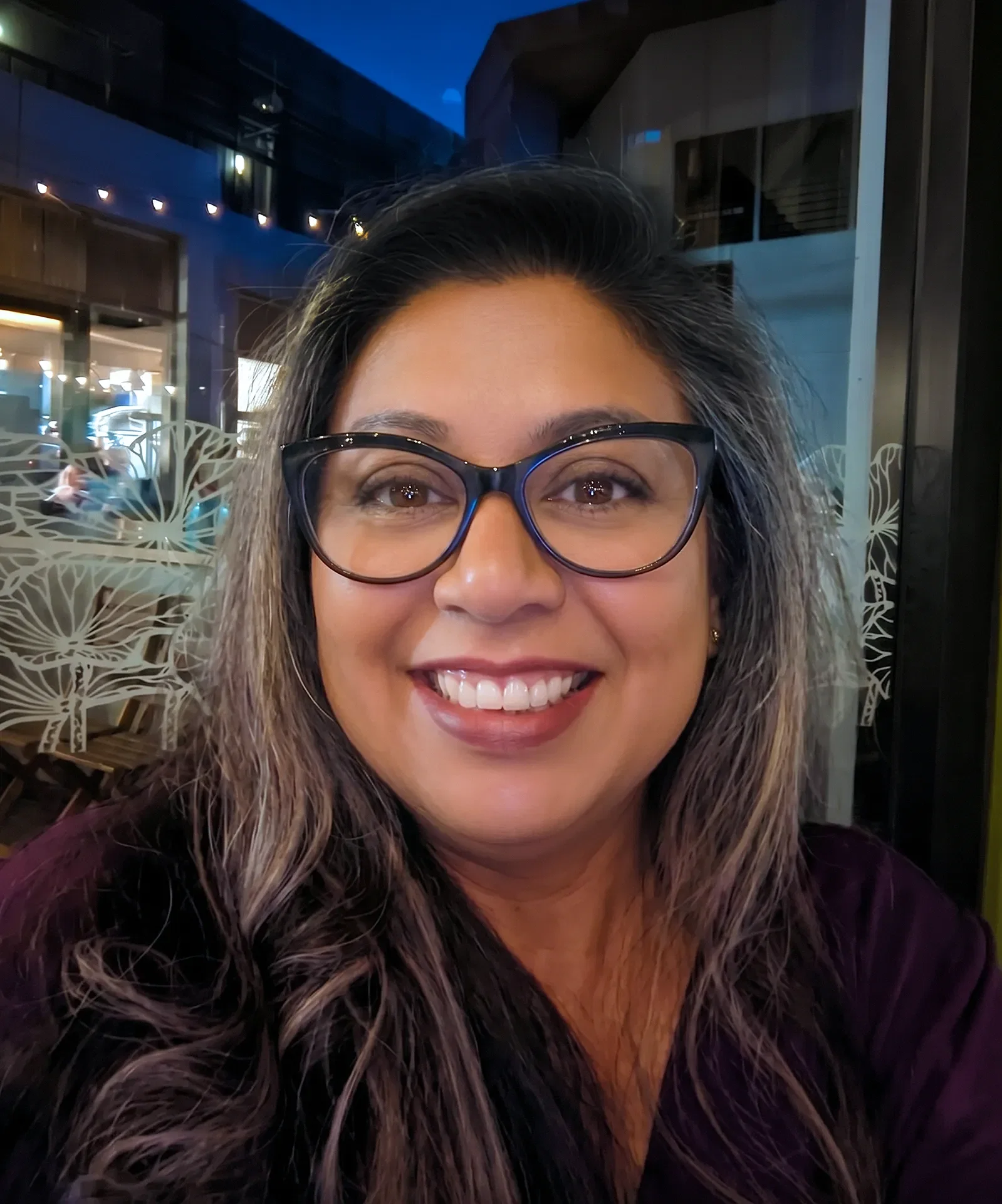 A woman with long dark hair and glasses smiles at the camera indoors at night. Decorative patterns on the window and San Francisco Bay Area lights outside create a warm ambiance.