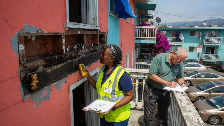 Two inspectors conduct an SB 326 balcony inspection on a pink apartment building; one uses a handheld device near a burnt section, while the other takes notes on a clipboard. Cars and other apartments are visible in the background.